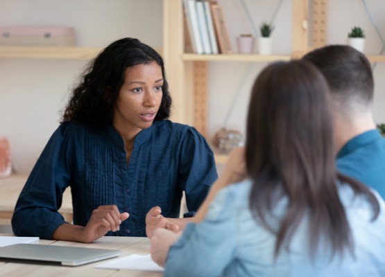 Focused African American female real estate agent or architect talk with young couple at meeting, concentrated biracial woman consultant speak discuss ideas with clients spouses at office briefing