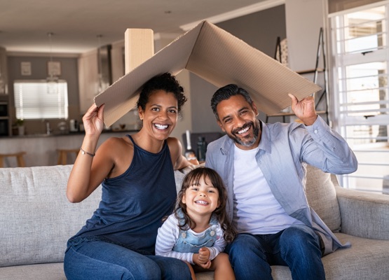 Portrait of smiling family sitting on couch holding cardboard roof and looking at camera. African and indian parents with funny daughter holding cardboard roof over heads while sitting on sofa in their new home. Happy mother and father with daughter in new house at moving day with copy space.
