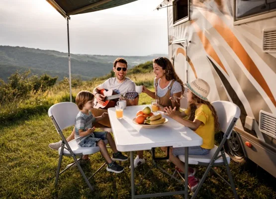 Happy family having fun while singing at picnic table by the camper trailer in nature. Man is playing a guitar.