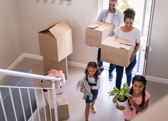 Ethnic family with two children carrying boxes and plant in new home on moving day. High angle view of happy smiling daughters helping mother and father with cardboard boxes in new house