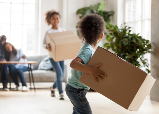 Happy African American family moving in new house, toddler boy carrying cardboard box, playing with preschooler sister, mother and father sitting on couch, looking at children, rear view, close up