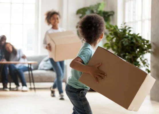 Happy African American family moving in new house, toddler boy carrying cardboard box, playing with preschooler sister, mother and father sitting on couch, looking at children, rear view, close up